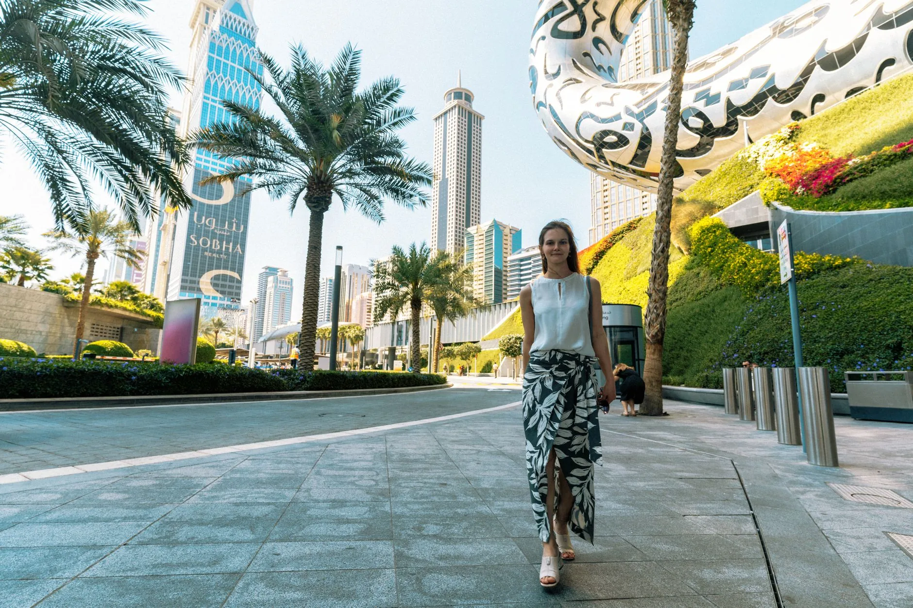 A woman walks in front of Dubai's iconic cityscape featuring towering skyscrapers and lush greenery.